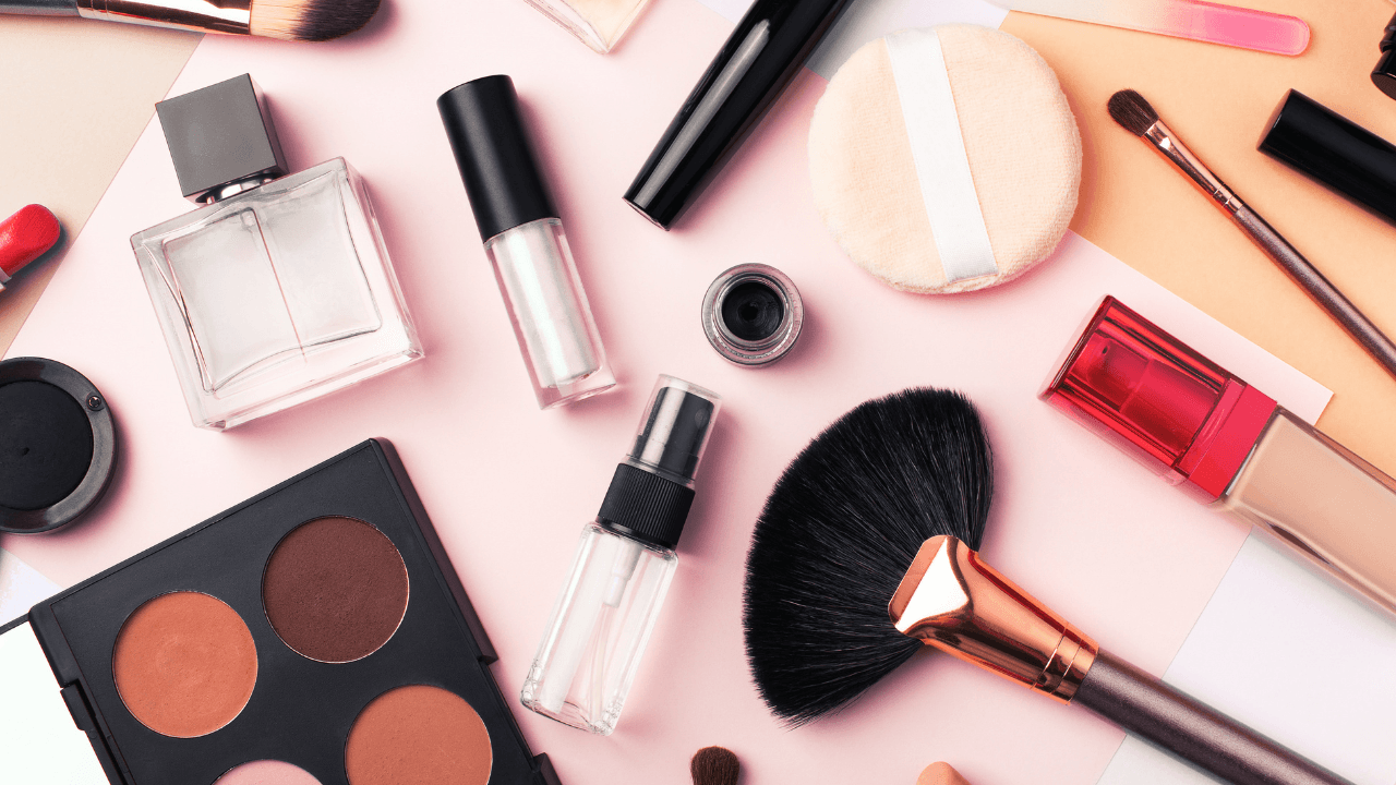 An editorial flat lay of brushes, palettes, and glass fragrance on blush pink.
